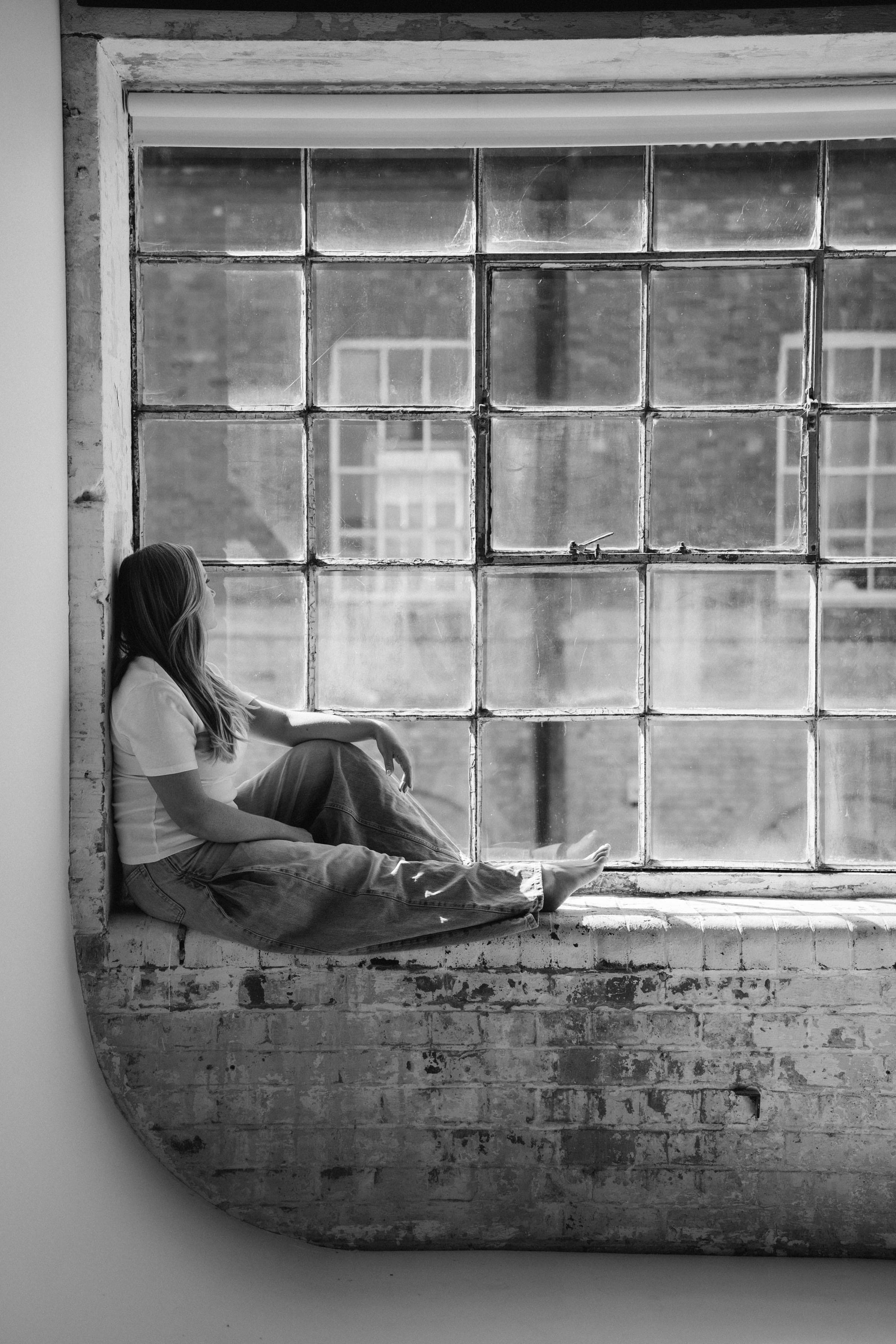 A woman sitting on the sill of a huge industrial window, looking out, black and white.