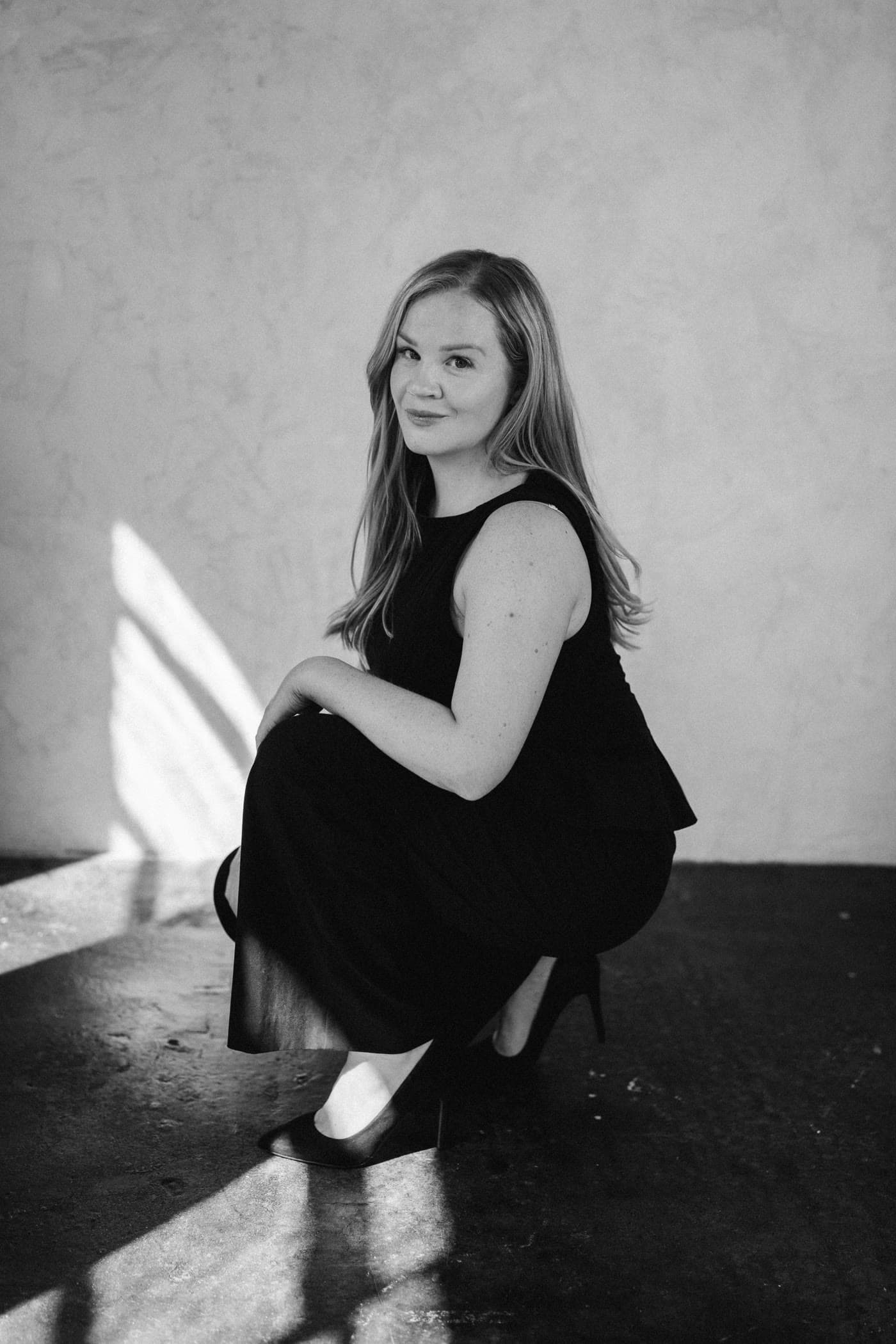 Grace, founder of Held By Grace, crouching in window light and smiling at the camera, black and white.